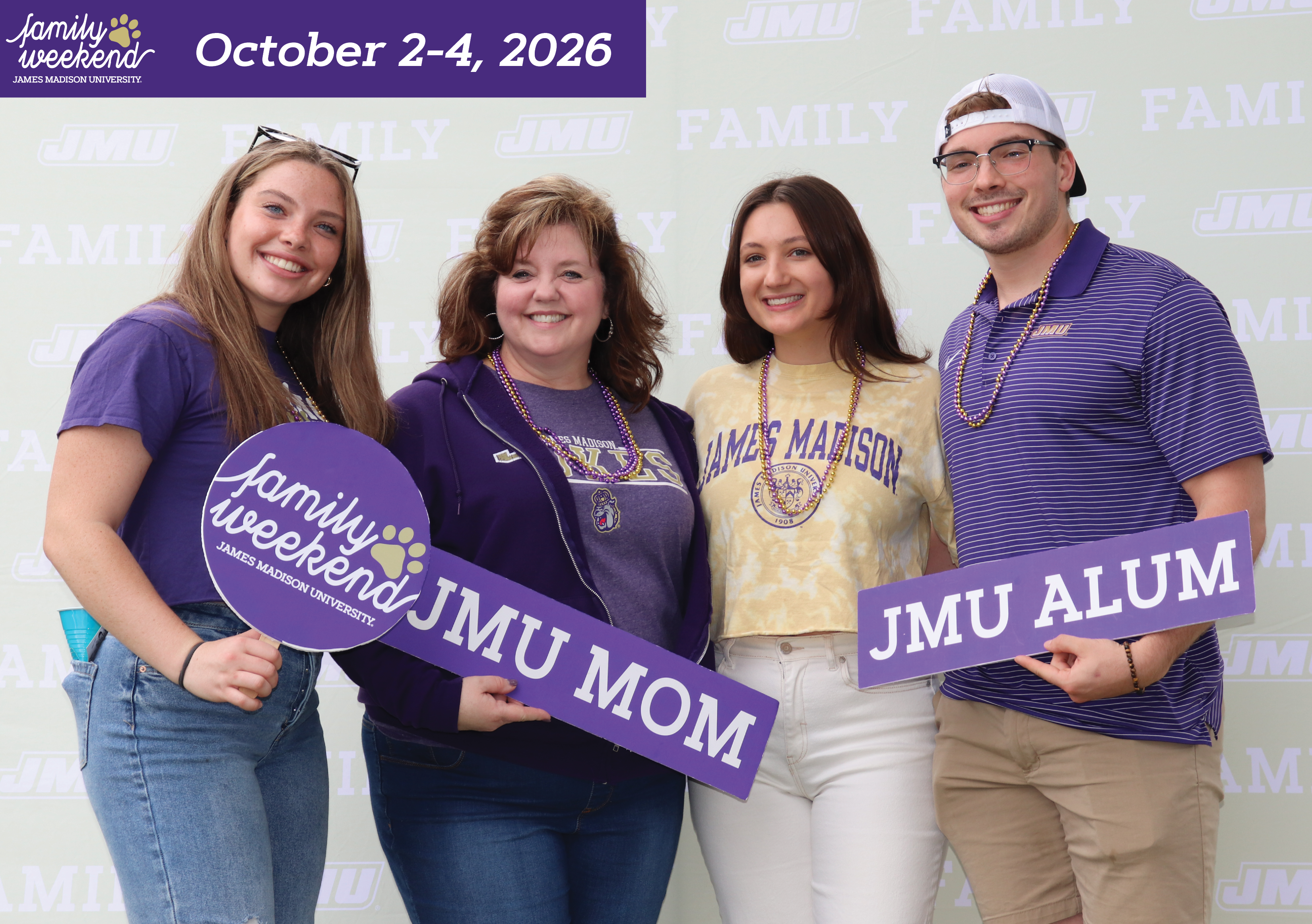A group of 4 JMU fans smiling for a photo dressed in JMU gear holding signs with their affiliation to the university. 